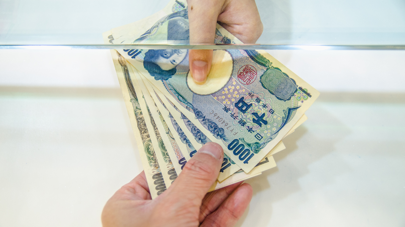 A person handing over a stack of Japanese yen banknotes through a glass counter, depicting a typical bank transaction in Japan.