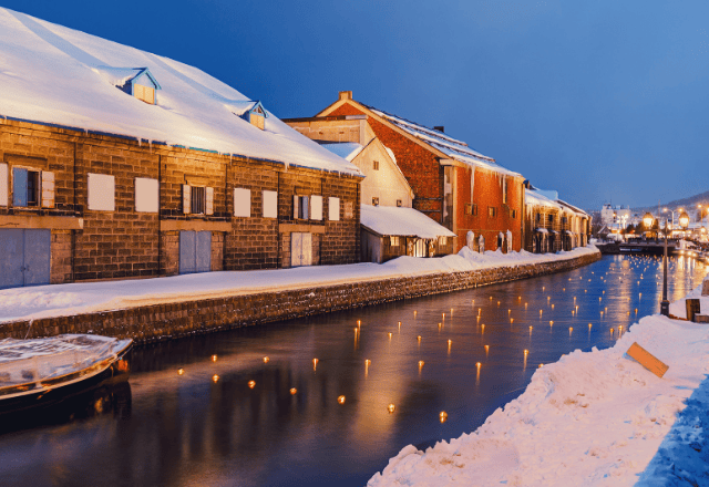 Winter night view of the Otaru Canal in Hokkaido, Japan, with snow-covered warehouses and illuminated lights along the water.