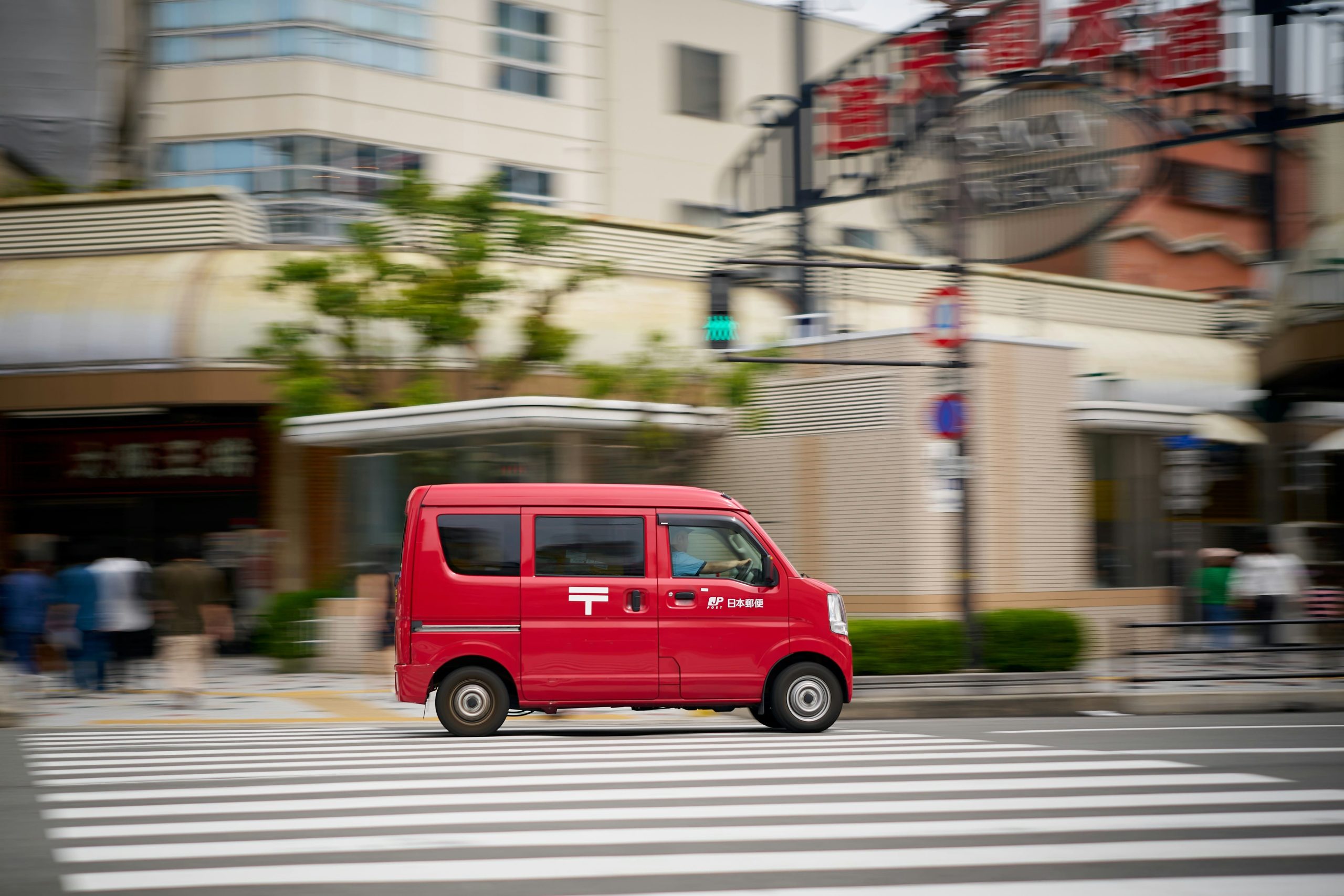 A red Japan Post van driving through a city street, symbolizing Japan's efficient postal delivery service.